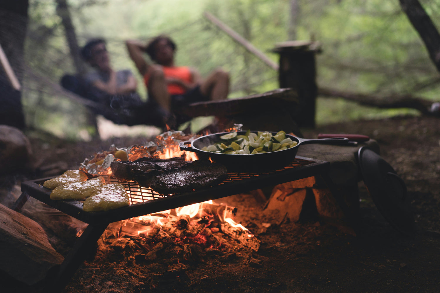 Outdoor cooking setup with a grill over an open fire, featuring a skillet of vegetables and sizzling BBQ food, perfect for camping.